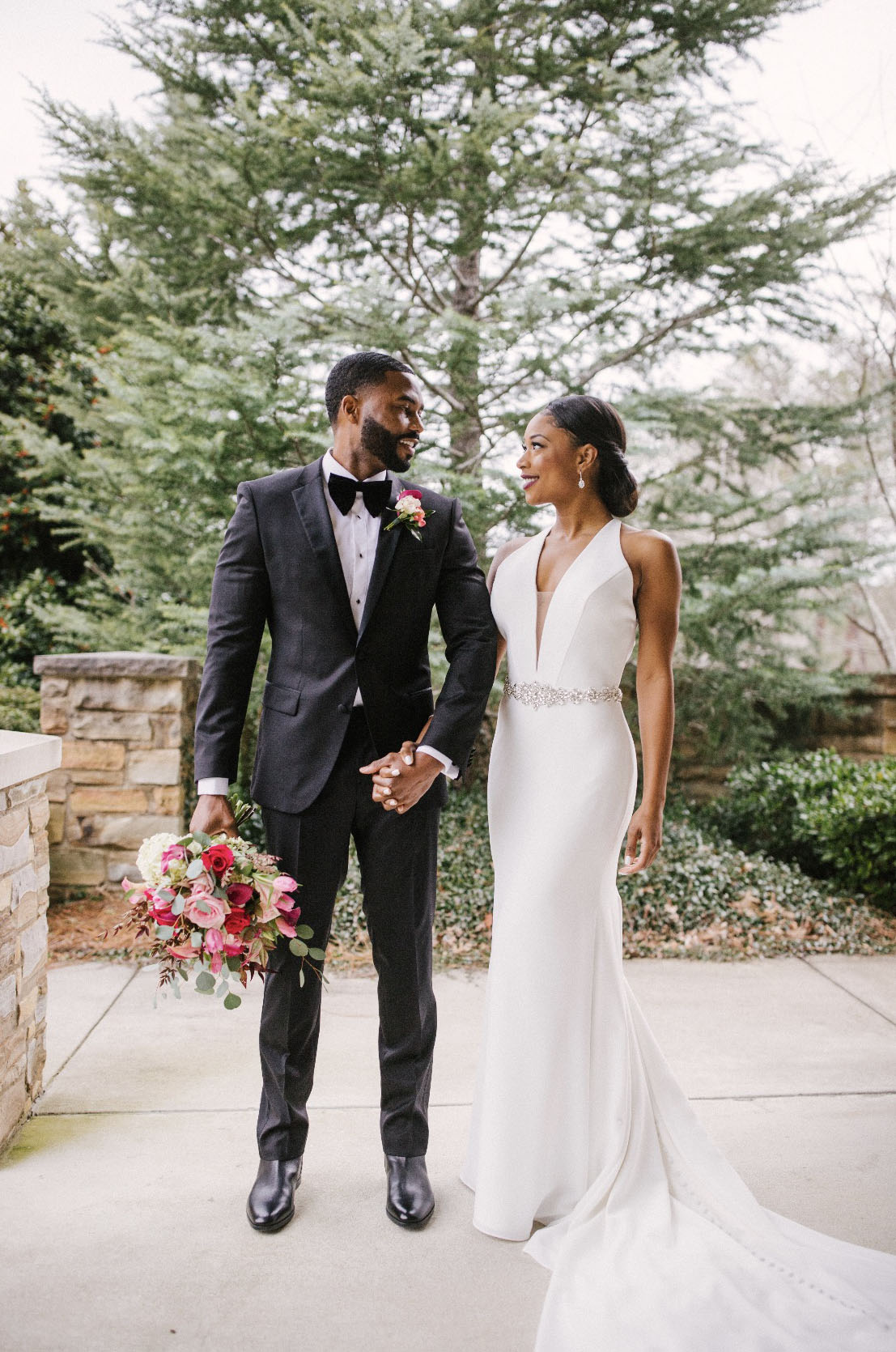 Bride in a plunging neck halter wedding dress with a belt and a crepe fit and flare skirt, poses with her groom on a terrace.