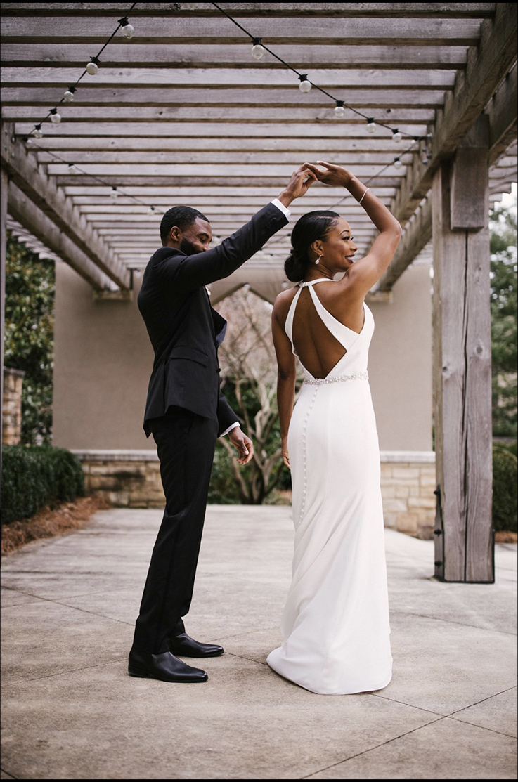 Groom twirls his bride in a keyhole back crepe gown with a buttoned skirt and a beaded belt, under a wooden pergola. 
