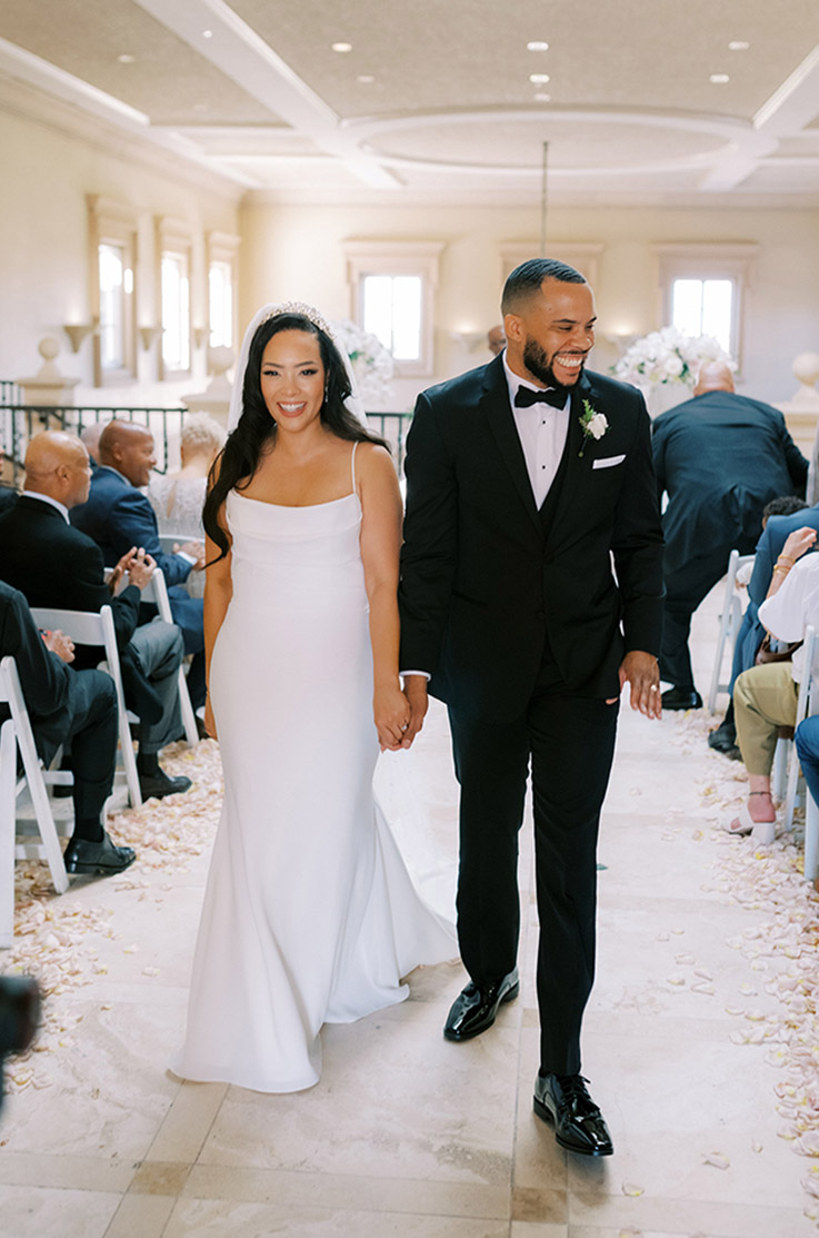 Bride in a crepe fit and flare gown with a scoop neck, a hairpiece and veil, walks hand in hand with her groom down the aisle.