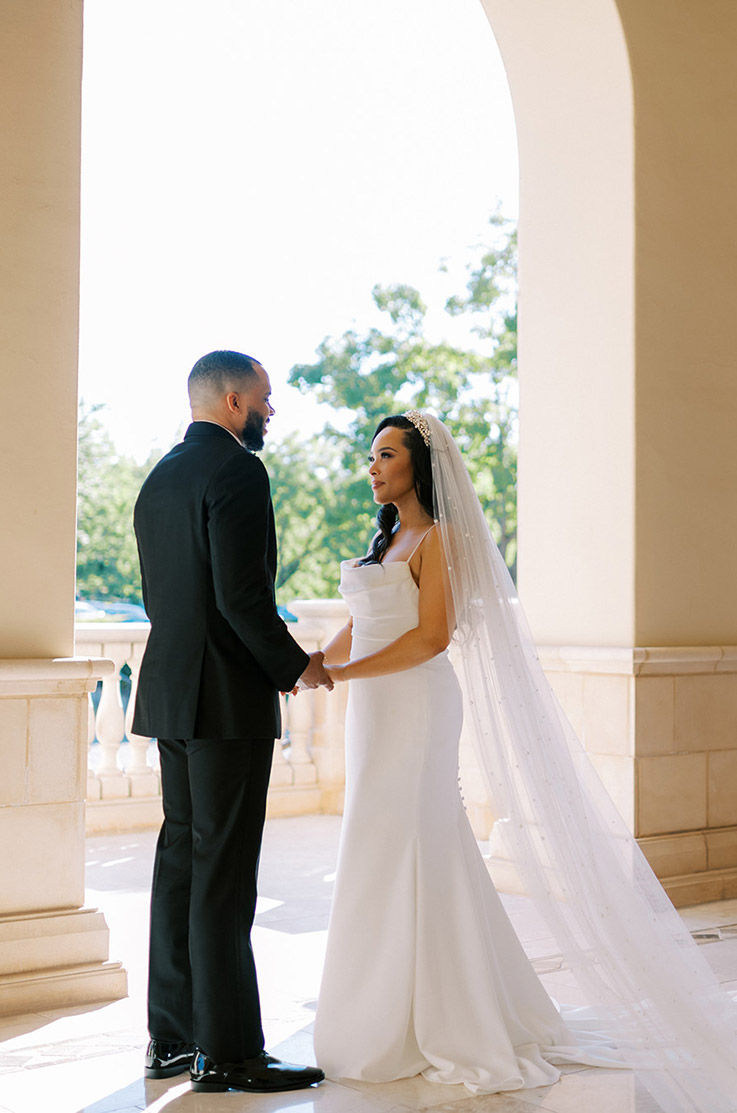 Bride in a scoop neck crepe gown with pleating, a buttoned skirt and a pearl veil, is facing her groom under a stone arch.