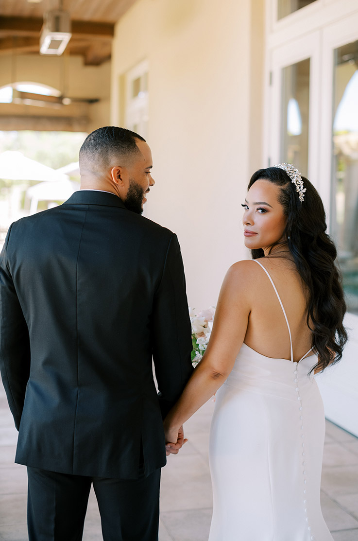 Bride looks past her shoulder in an open back crepe gown with buttons, walking hand in hand with groom outside the venue.