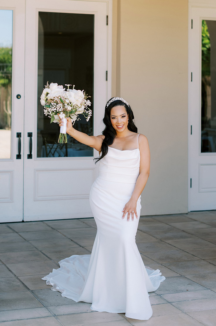 Bride in a scoop neck crepe gown with a fit and flare skirt and pleating, holds a bouquet up outside venue doors on a patio.