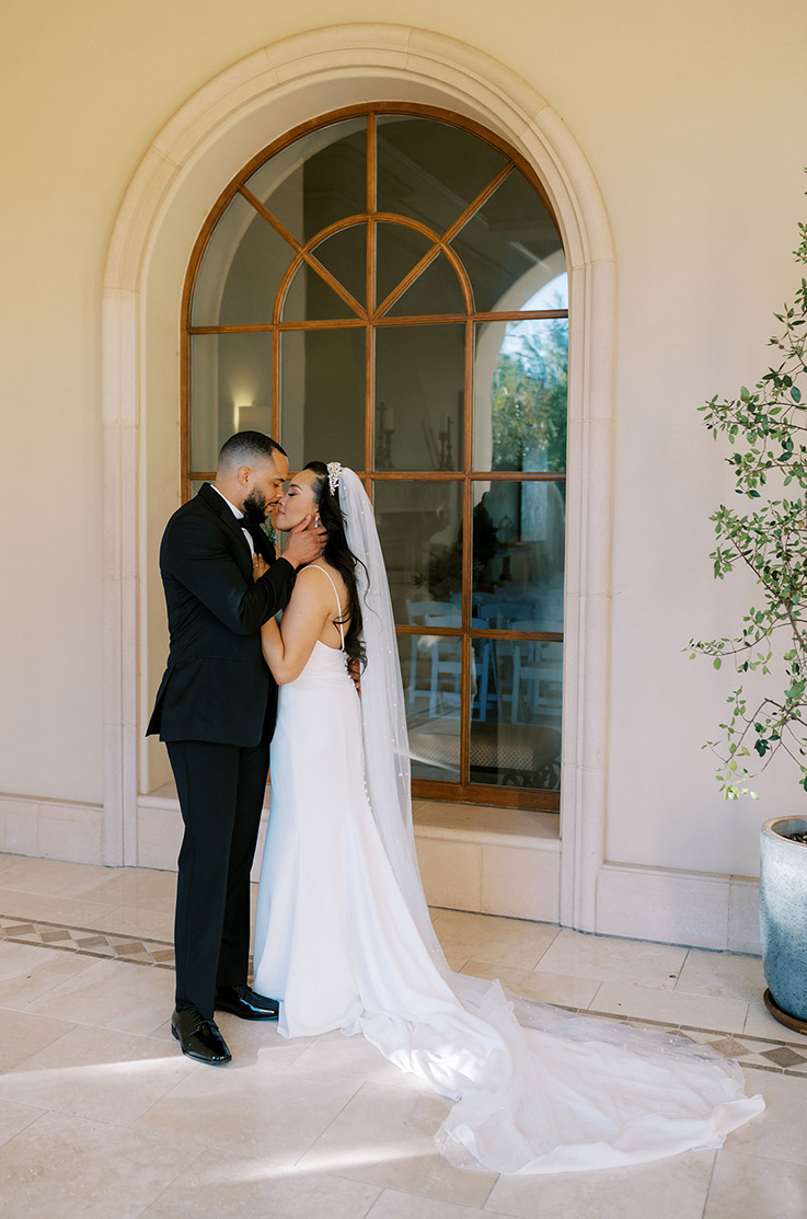 Groom kisses his bride in a low back crepe wedding dress with a fit and flare skirt and veil, in front of a tall arch window.