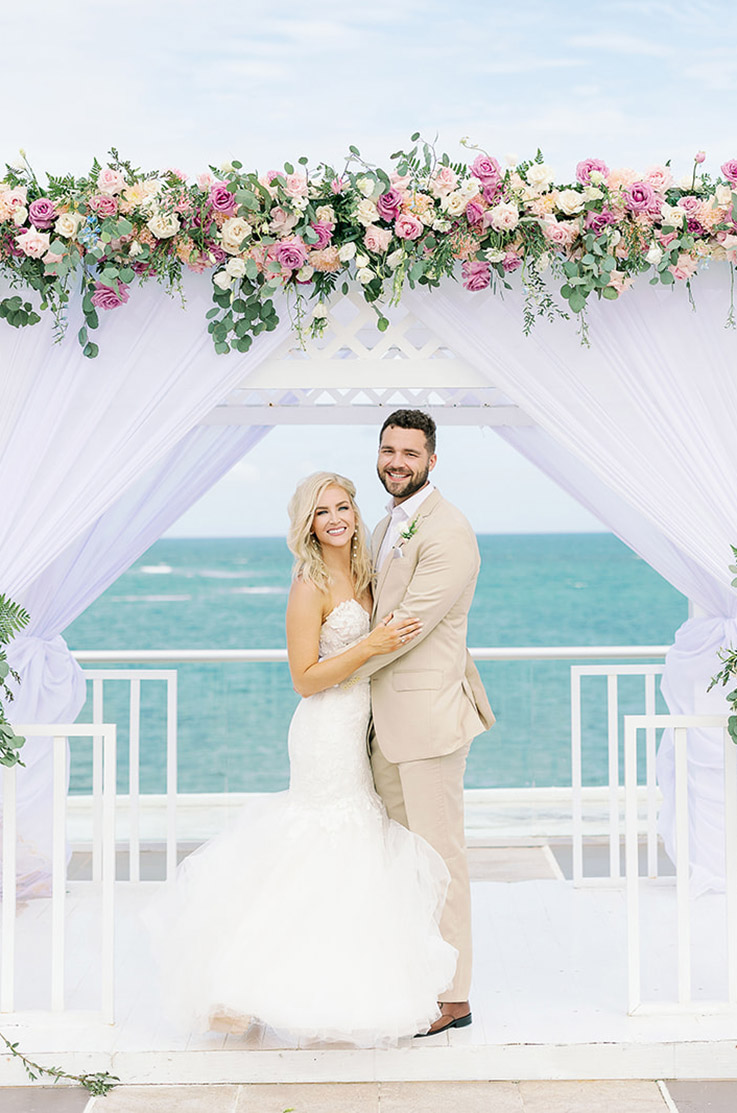 Groom embraces his bride in a tulle mermaid gown with a strapless lace bodice, under a floral arbor at an oceanside.