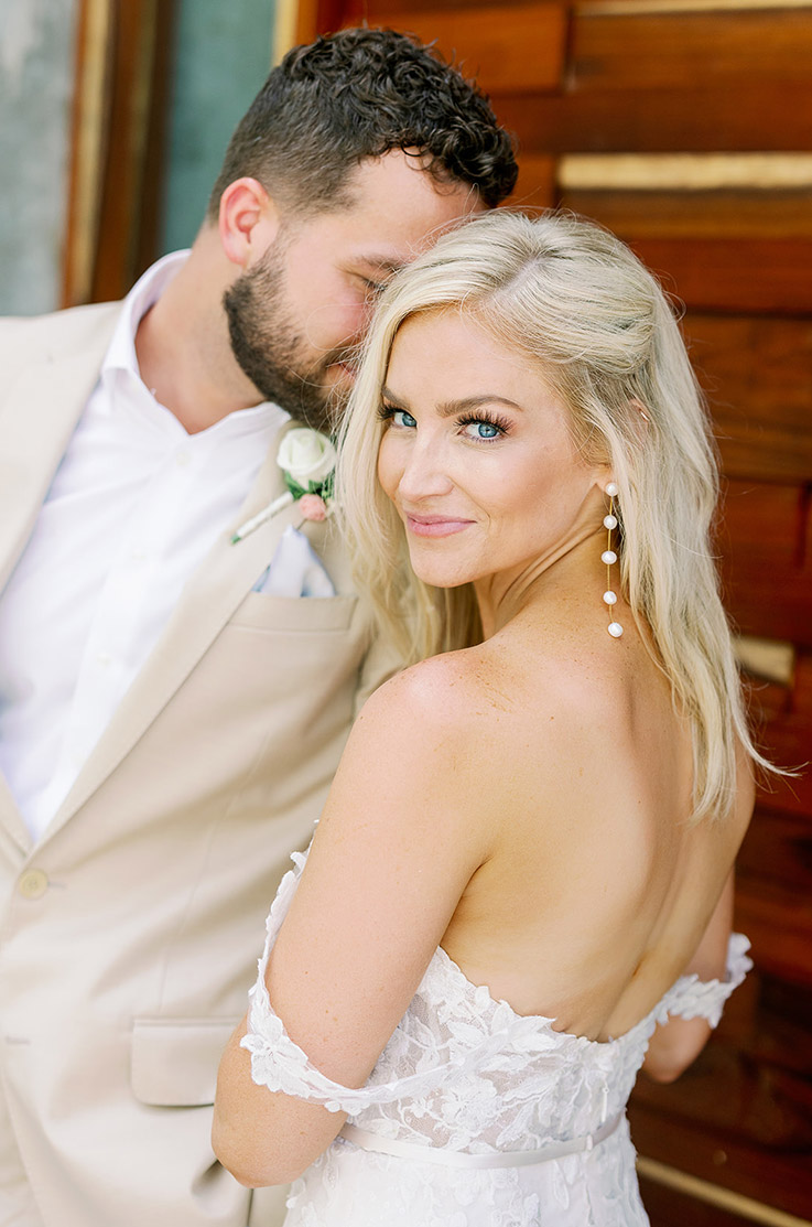 Close-up of bride in a low back lace gown, lined in tulle with a thin fabric belt, beside the groom in a tan suit.