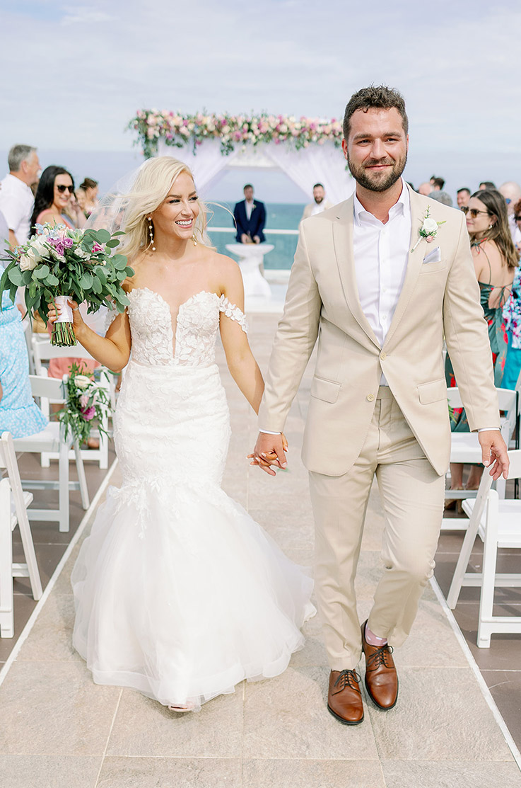 Bride in an off-shoulder lace and tulle gown, is holding a bouquet and walks with groom down the aisle by an oceanfront.