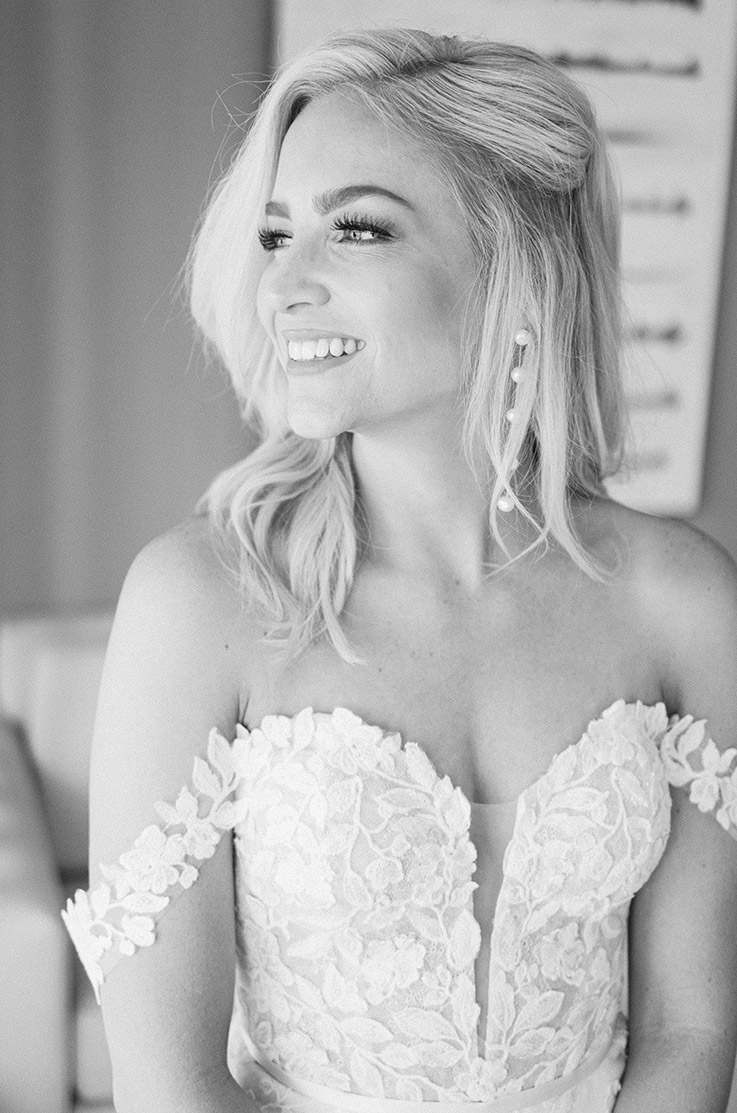 Close-up of bride smiling in a plunging neck strapless gown with lace appliques on the bodice, indoors in black and white.
