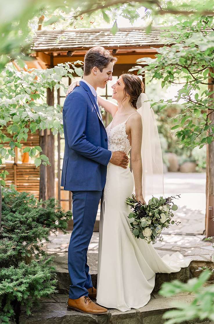 Bride in a lace bodice gown with a fit and flare crepe skirt and a veil, looks up at her groom at a garden pavilion.