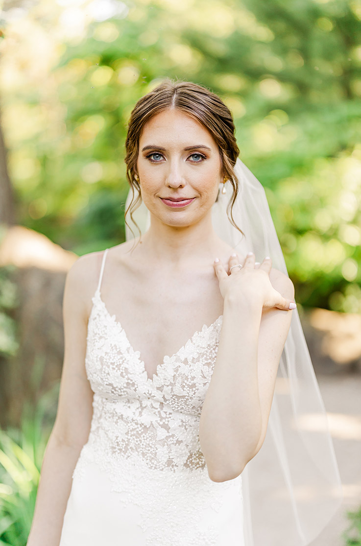 Close-up of bride in a V-neck lace bodice gown with spaghetti straps and a veil, poses in a lush garden.