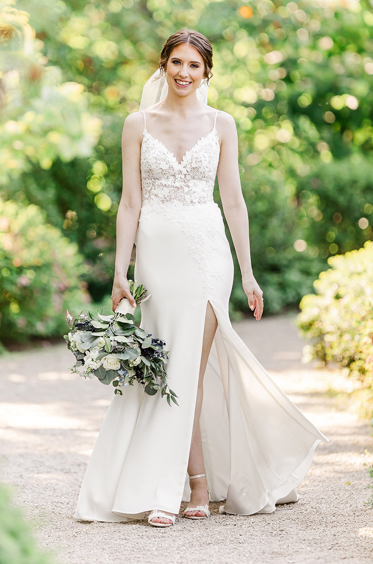 Bride stands on a garden path in a lace and crepe gown with a V-neckline, fit and flare skirt and a veil, holding a bouquet.