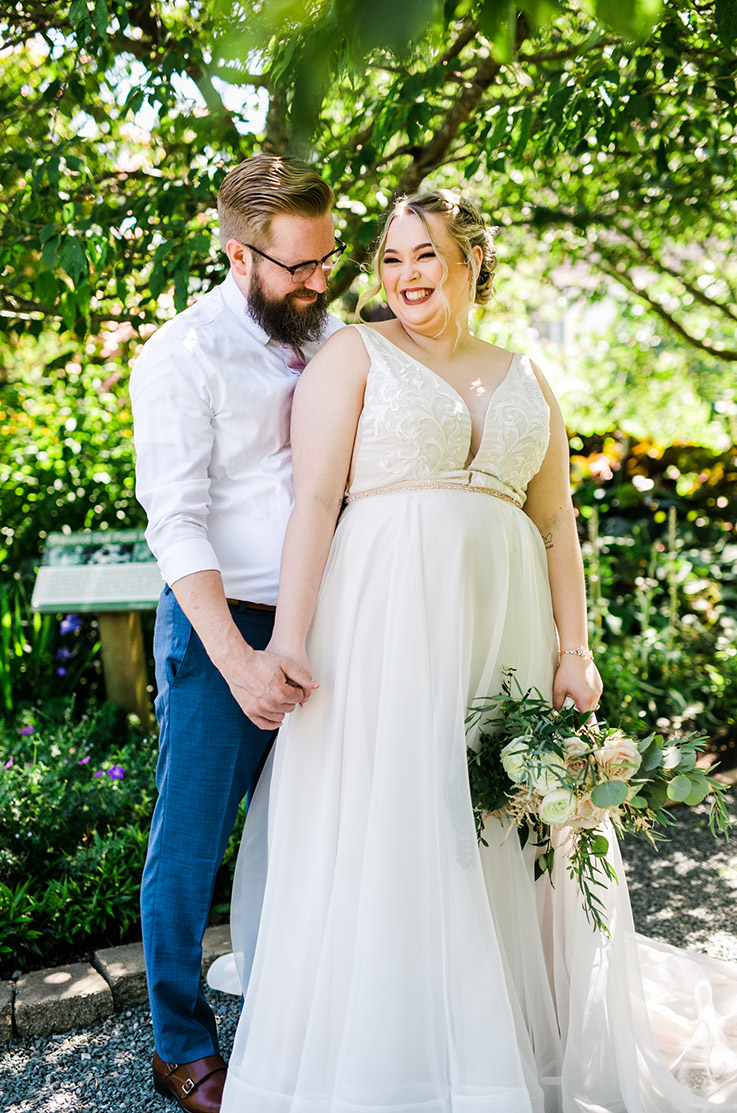 Bride in a plunging V-neck gown with embroidered appliqueing and an organza skirt, holds a bouquet in a sunny outdoor area. 