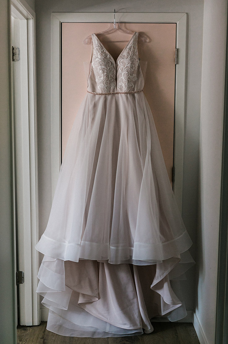 A V-neck embroidered applique gown with belt detail and an organza skirt, hangs on a doorway indoors.