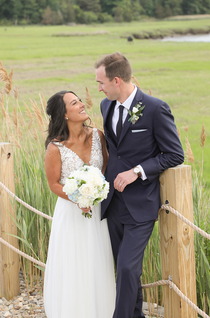 Bride in a V-neck tulle gown with lace detail, holds a bouquet and poses beside her groom at a wooden fence and tall grass.