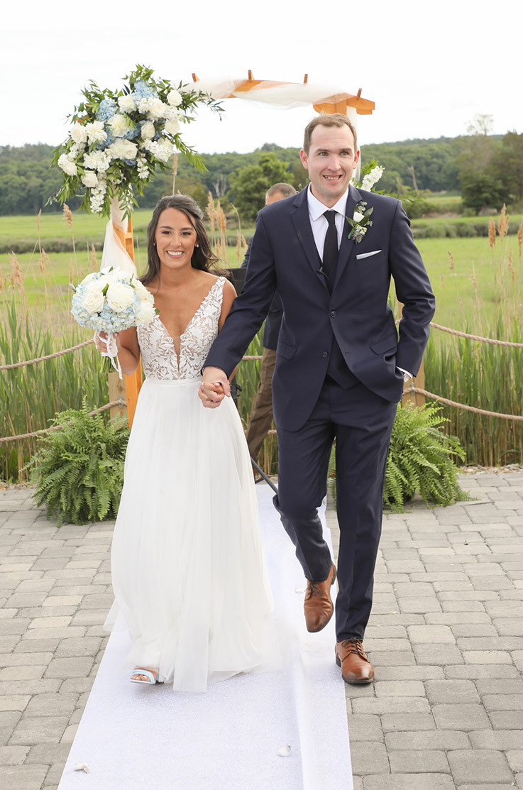 Bride in a sleeveless V-neck gown with a tulle skirt and lace appliques on the bodice, walks alongside her groom in the aisle.