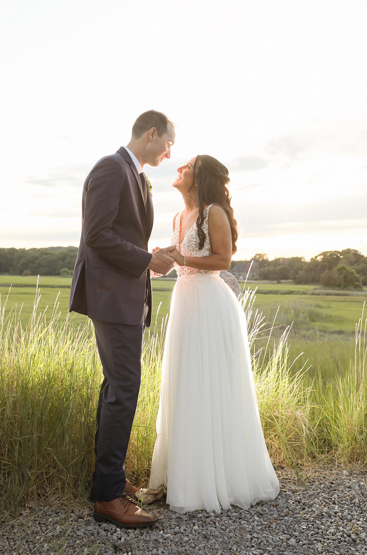Bride in a V-neck lace bodice gown, lined in tulle with a full skirt, looks up at her groom in a sunny open meadow. 