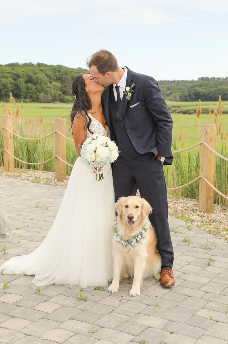 Groom kisses bride in a lace bodice wedding dress with a flowing tulle skirt, holding a bouquet beside their dog at a meadow.