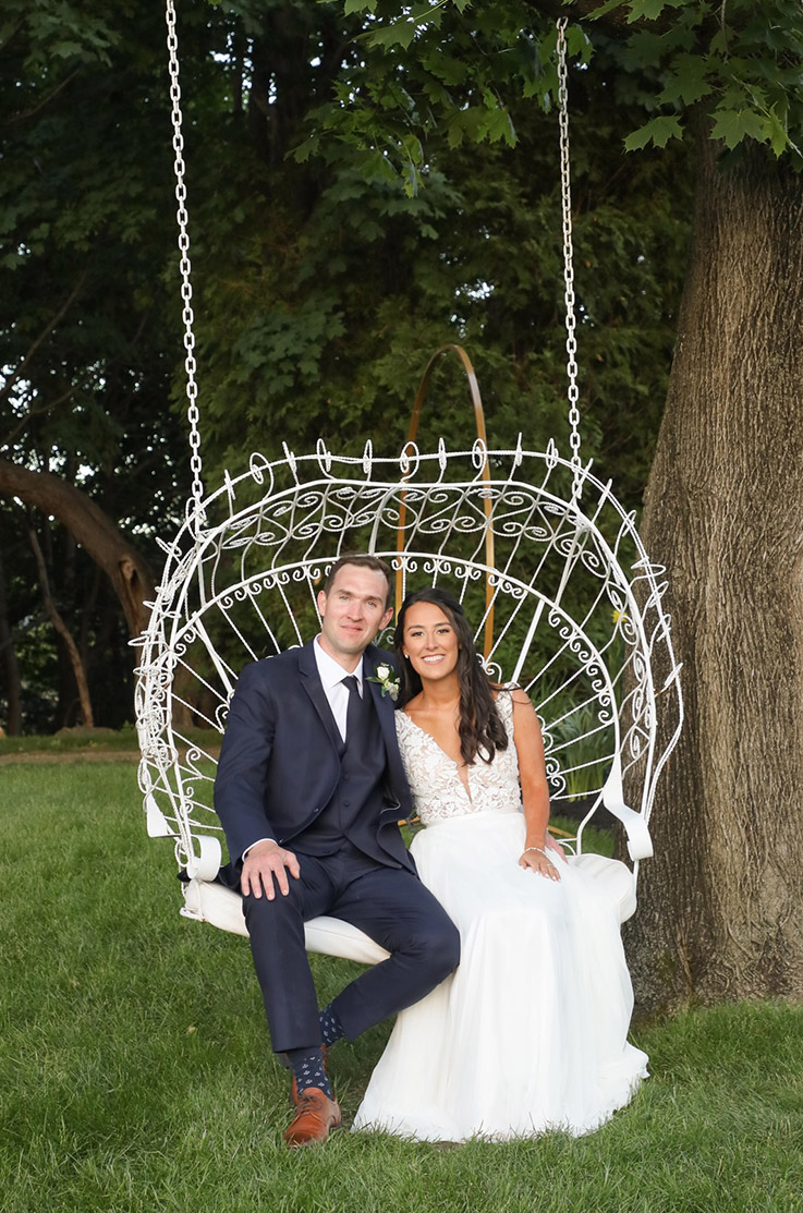 Groom sits next to bride in an outdoor swing, in a V-neck lace and tulle gown with a flowing skirt, under a large tree.
