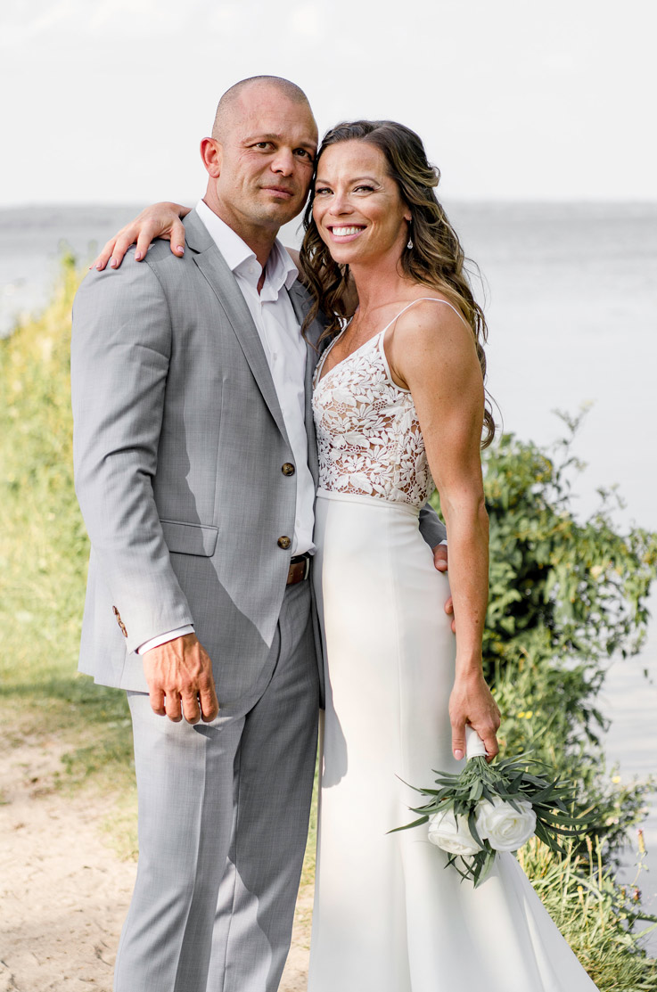 A bride in a V-neck wedding dress, holding a bouquet, and a groom in a suit hold each other while standing outdoors in front of a lake and smiling.