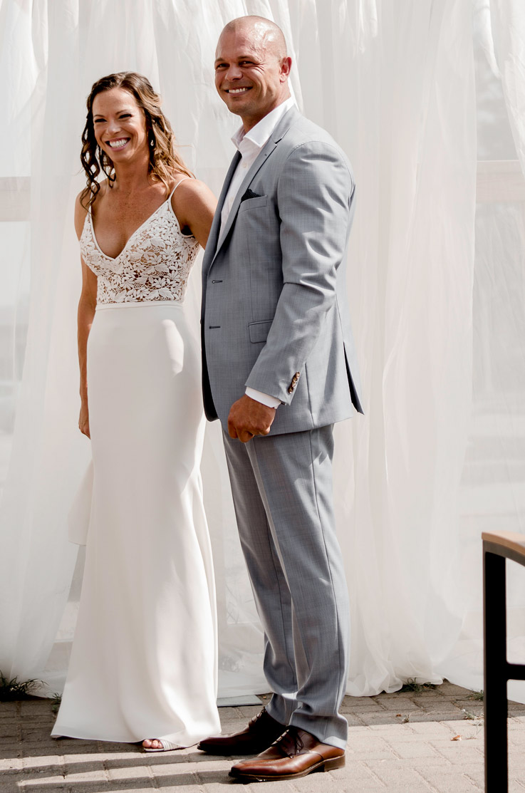 A bride in a V-neck wedding dress and a groom in a suit stand together and smile outdoors, in front of flowing curtains on a brick patio.