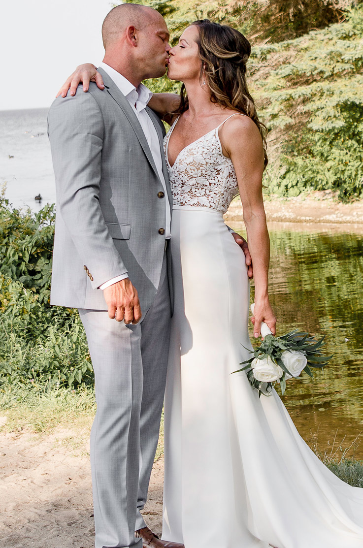 A bride in a V-neck wedding dress, holding a bouquet, and a groom in a suit hold each other and kiss while standing outdoors in front of a lake.