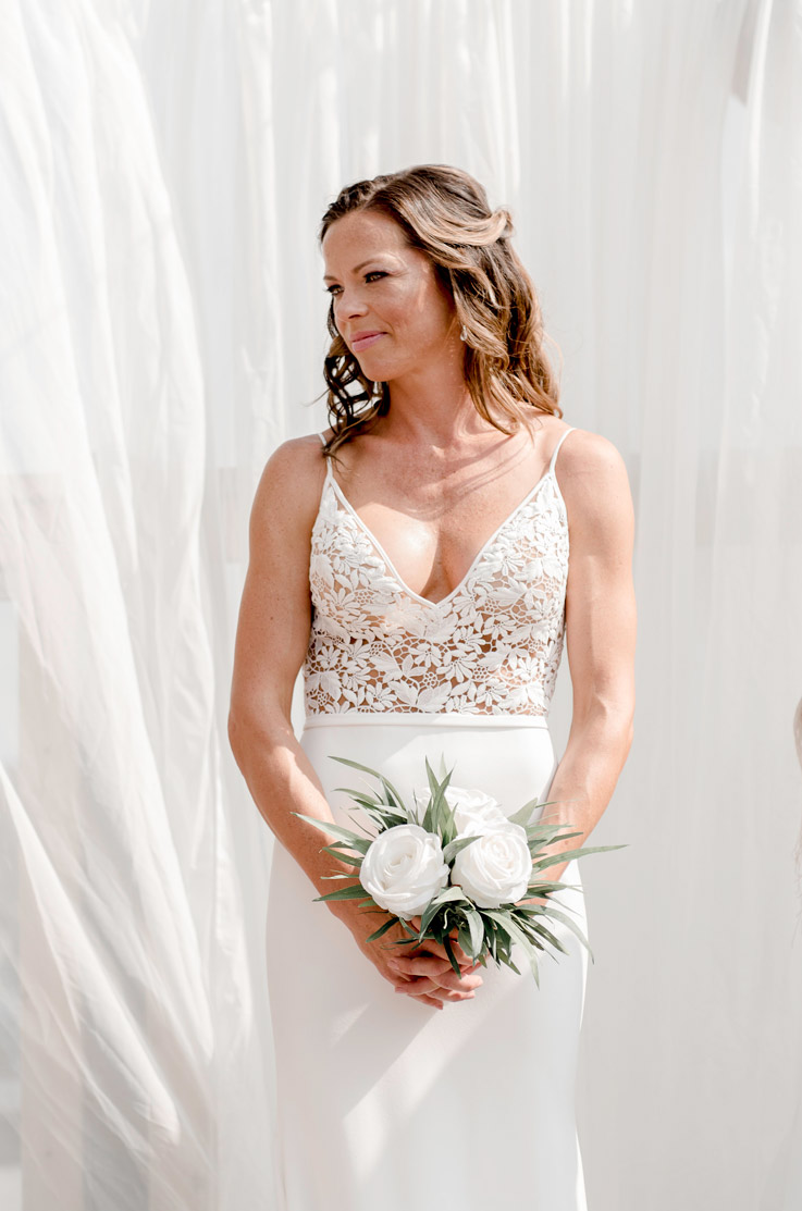 A bride in a V-neck wedding dress holds a bouquet while standing outside in front of flowing curtains.