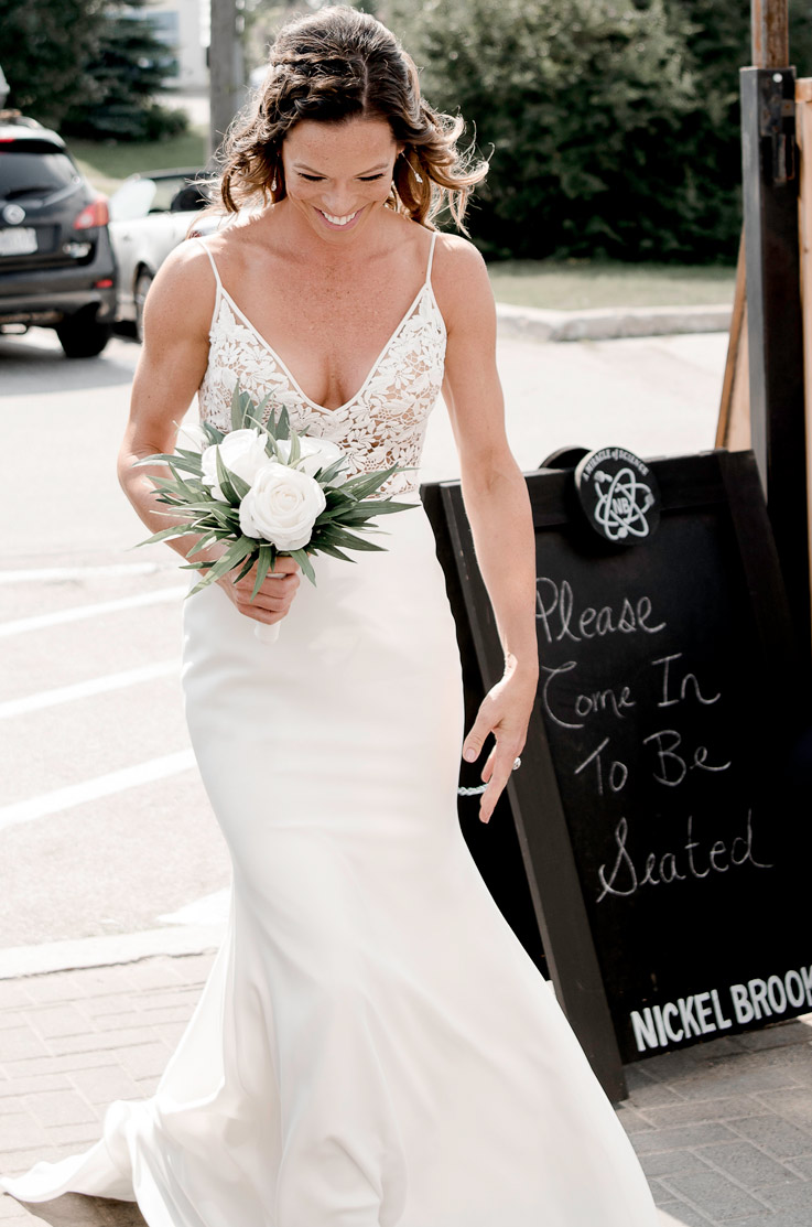 A bride in a V-neck wedding dress, holding a bouquet, walks down a sidewalk outside in a city.