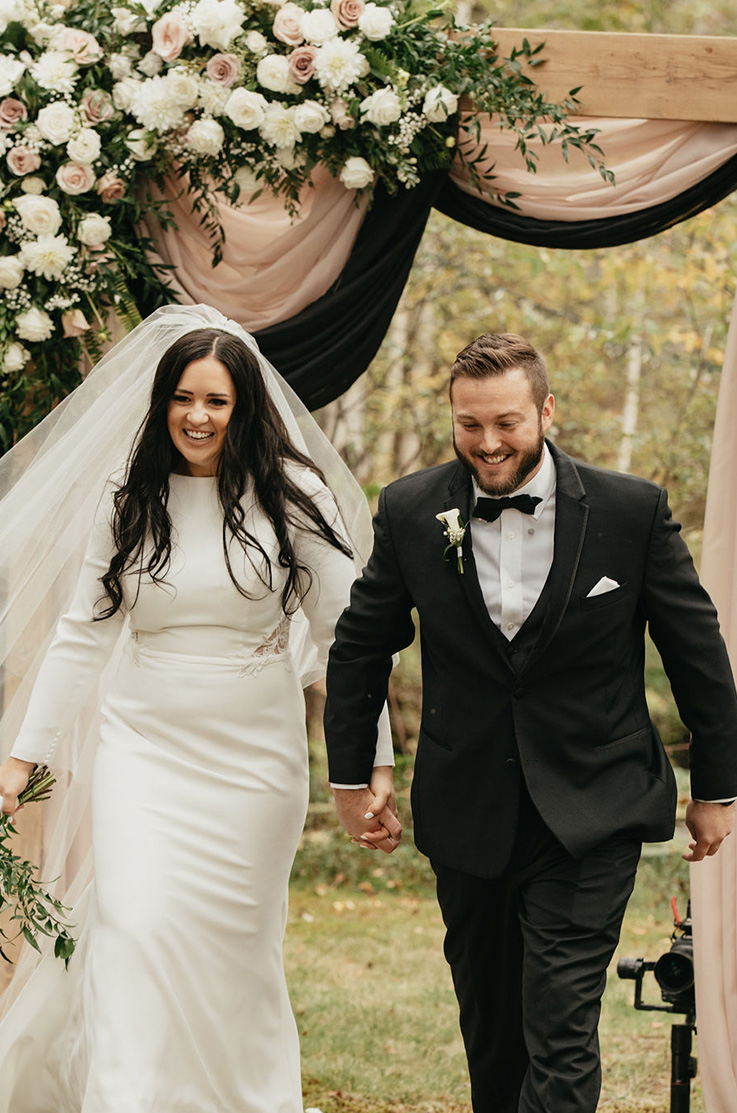 A bride in a long sleeve wedding dress and veil, and a groom in a suit, hold hands and smile in front of a decorated wedding arch outdoors, with trees in the background.