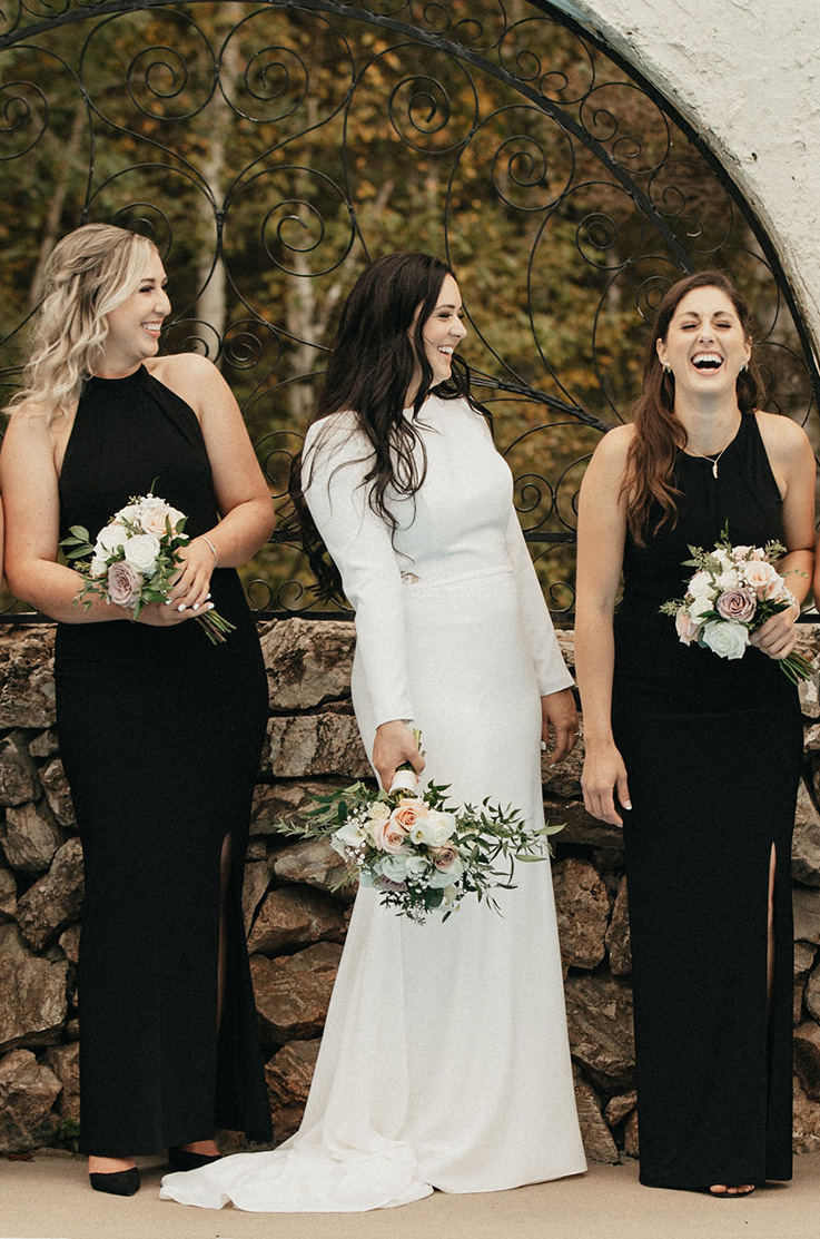 A bride in a long sleeve wedding dress stands outside with two bridesmaids, all of them holding bouquet and laughing while standing in front of a half stone wall and decorative iron bar window.