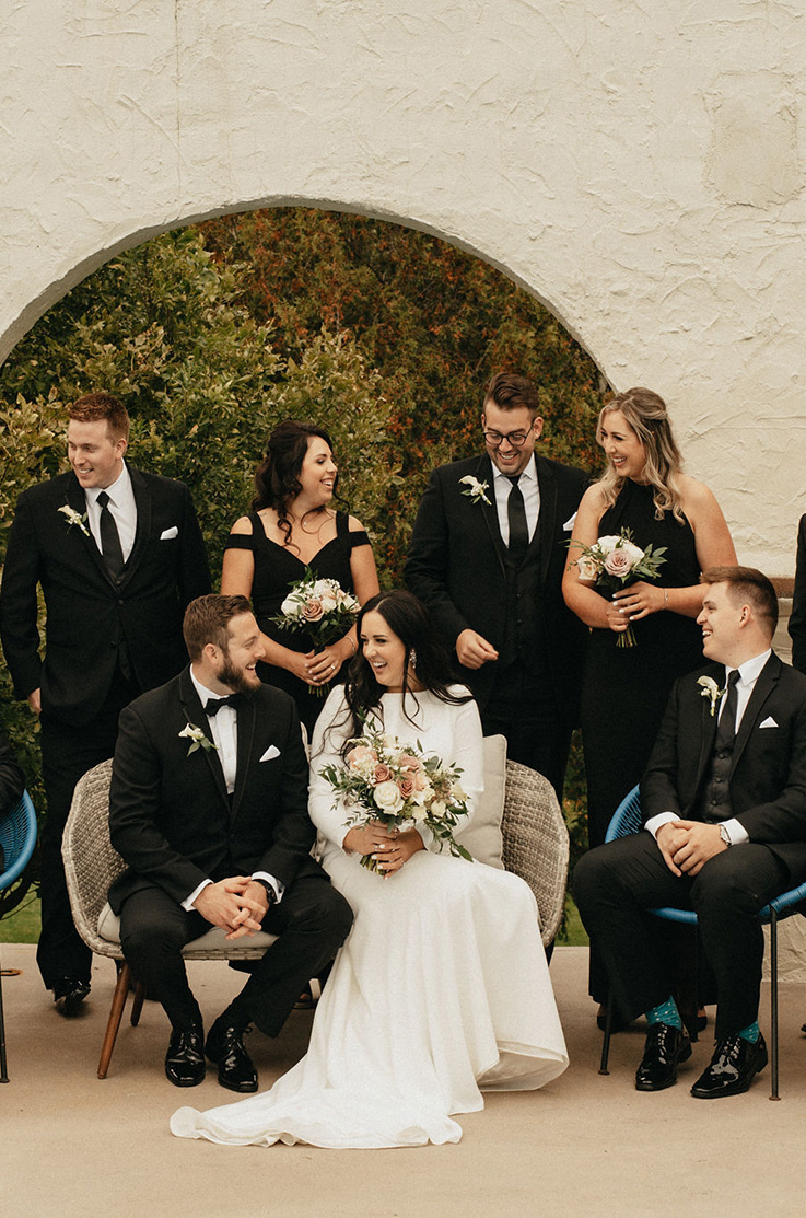 A bride in a long sleeve wedding dress and a groom in a suit sit on an outdoor loveseat on a patio, with their wedding party around them, all laughing and smiling.