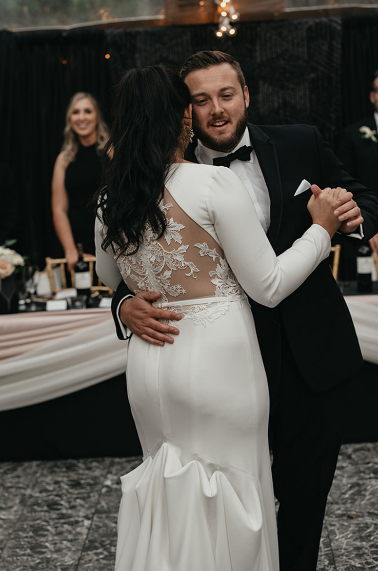 A bride in a lace back, bustled wedding dress, and a groom in a suit dance together, with the head table and a bridesmaid behind them.