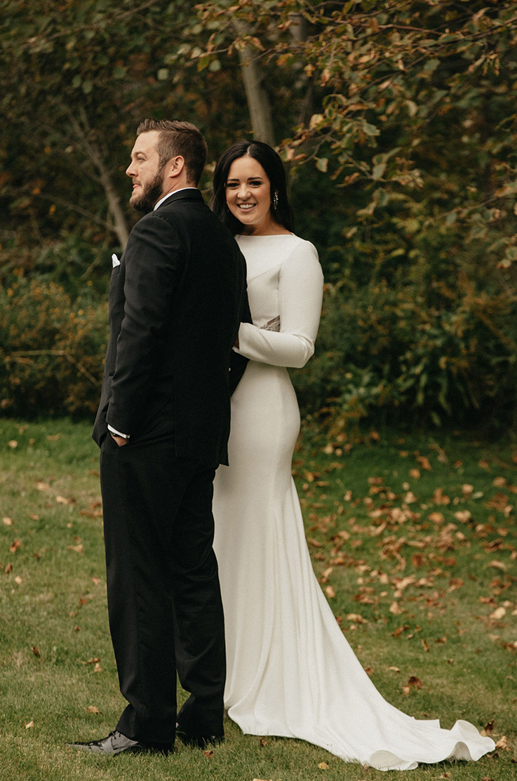 A smiling bride in a long sleeve wedding dress holds the arm of a groom in a suit while they stand outside in the grass, with leaves and trees in the background.