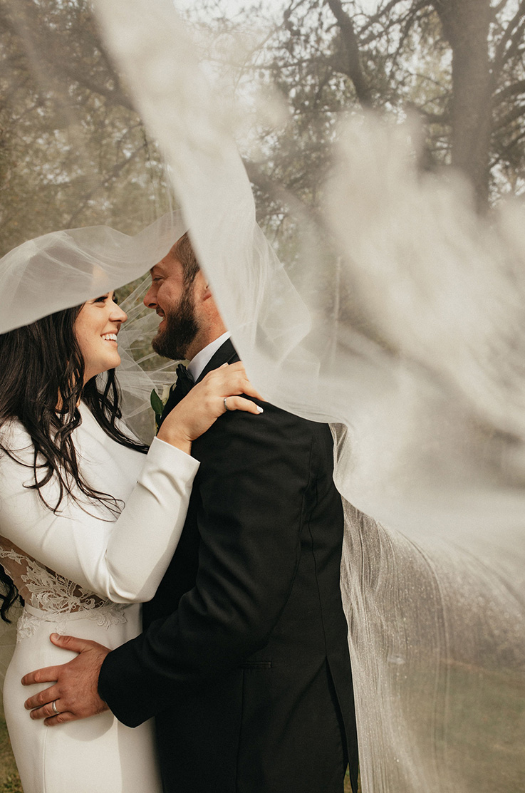 A bride in a long sleeve wedding dress and a groom in a suit hold each other and smile while looking into each others eyes, with the bride's veil draped over them.