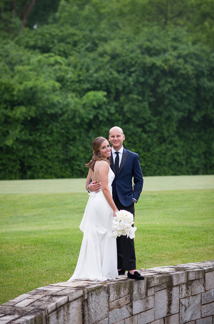 A bride in an embellished wedding dress, holding a bouquet, and a groom in a suit stand on a stone wall outdoors, with a grassy field and trees behind them.