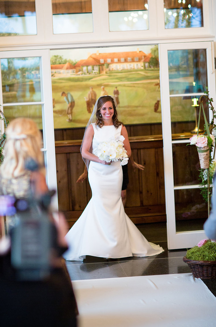 A bride in a sleeveless wedding dress holding a bouquet makes an entrance through open glass panel doors inside.