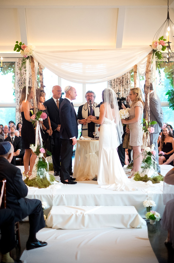 A bride in an embellished wedding dress and veil, and a groom in a suit, stand at an altar decorated with greenery with a Rabbi and their wedding party, as they say their vows. 