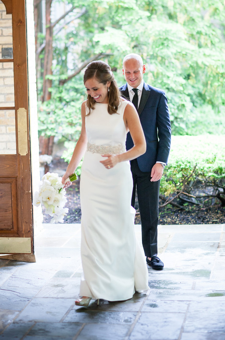 A groom in a suit admires a bride in an embellished wedding dress as she holds a bouquet, while they both walk into a building with a stone floor and large wooden doors.