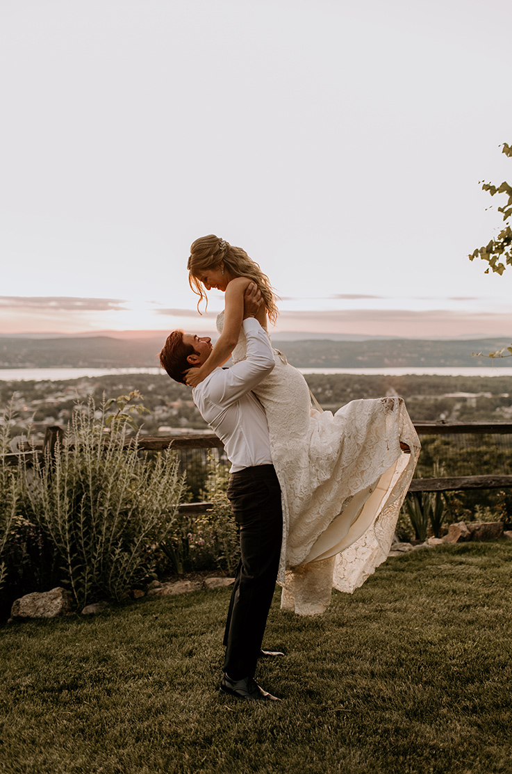 A groom in a dress shirt lifts a bride in a lace wedding dress in the air while standing in the grass with a wooden fence and a scenic view of a valley behind them.