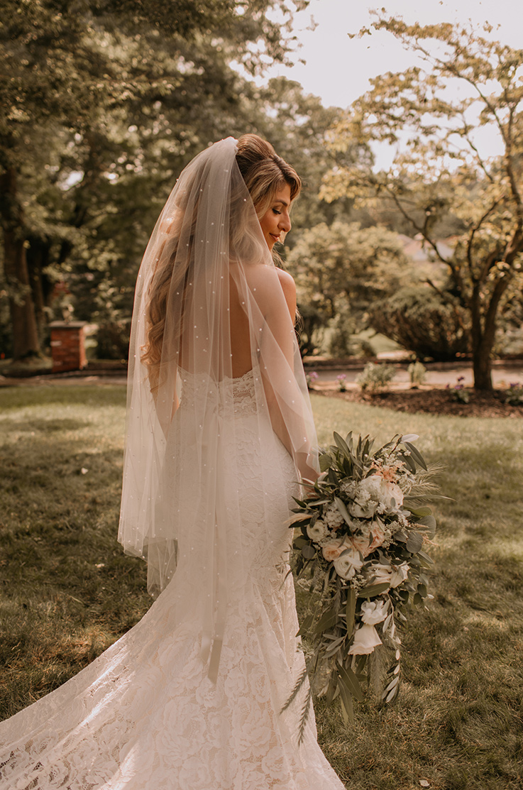 A smiling bride in a lace wedding dress and veil, holding a bouquet, stands outside in the grass with trees in the background.