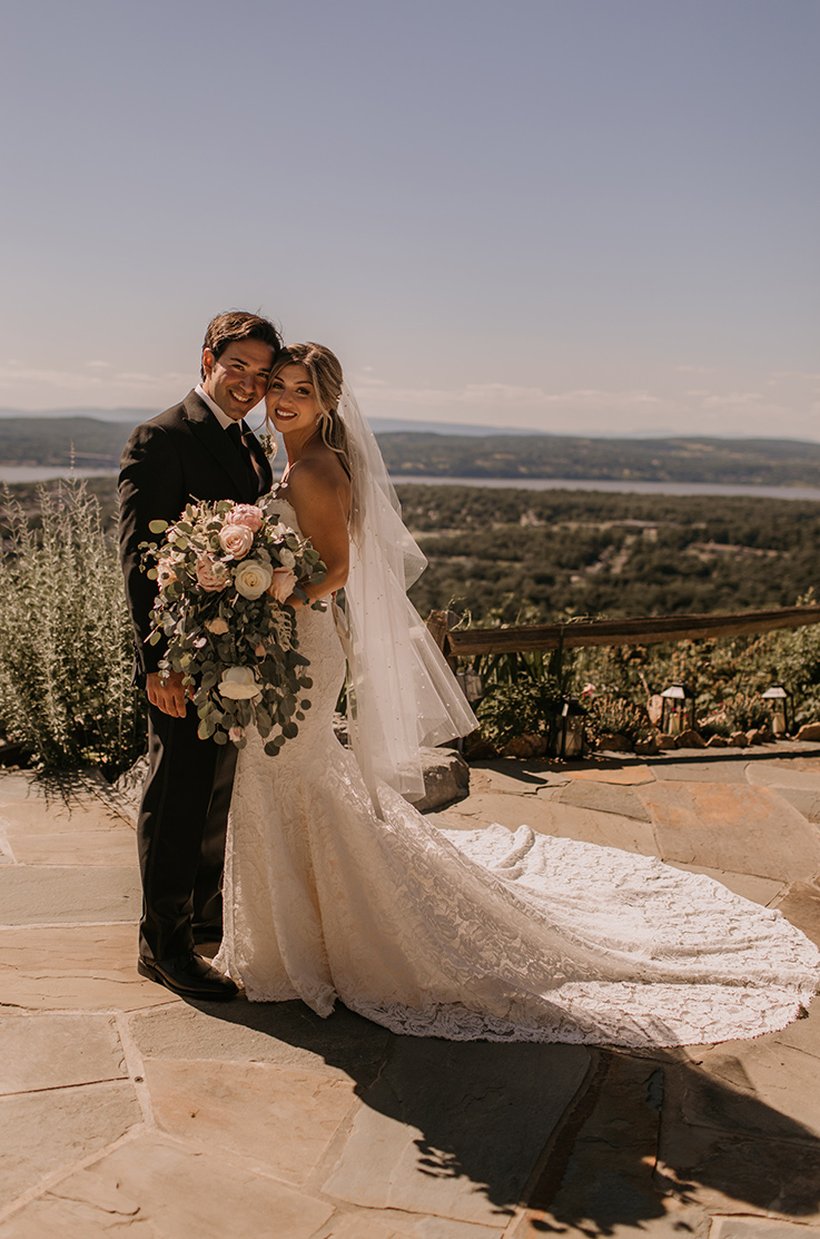 A bride in a lace wedding dress and veil, holding a bouquet, and a groom in a suit, stand on a stone balcony with a scenic view of a valley behind them.