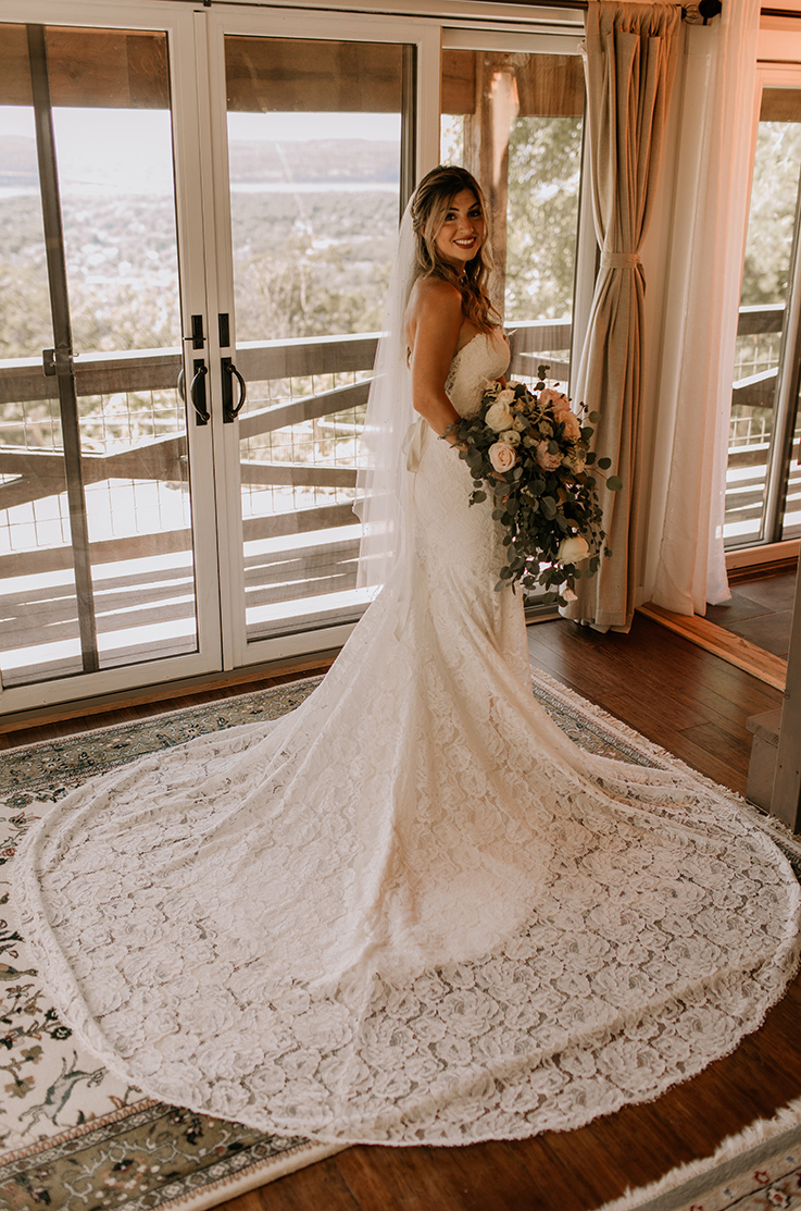 A smiling bride in a lace wedding dress and veil, holding a bouquet, poses indoors in front of a set of glass doors with her train fanned out to her side.