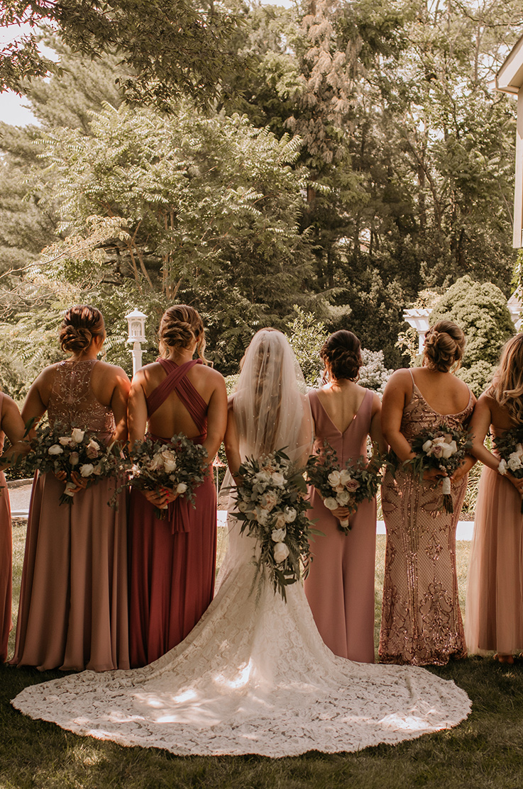 A bride in a lace wedding dress and veil, and 6 bridesmaids, face away and hold their bouquets behind their backs while standing in the shade outside, with trees and lamp posts in the background.
