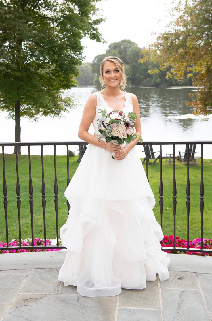 A bride in a tulle wedding dress holds a bouquet while standing on a balcony in front of a grass yard, lake, and trees.
