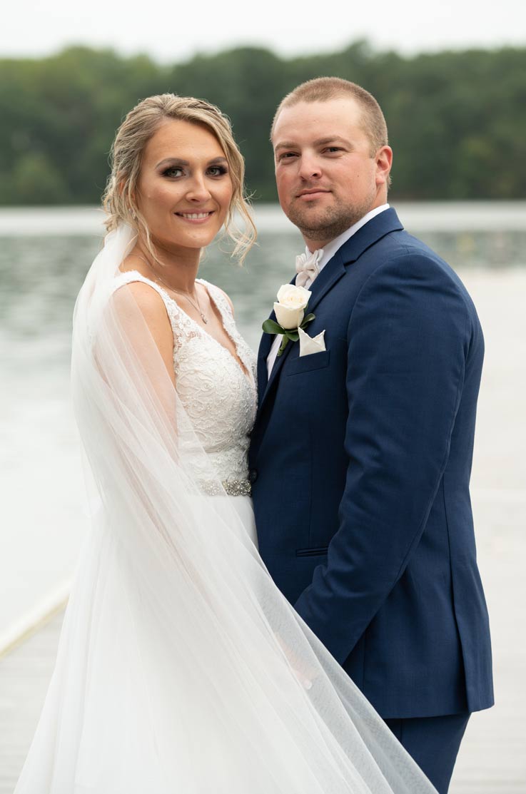 A bride in a sleeveless wedding dress and veil, and a groom in a suit, stand together outside in front of a lake, with trees in the background.