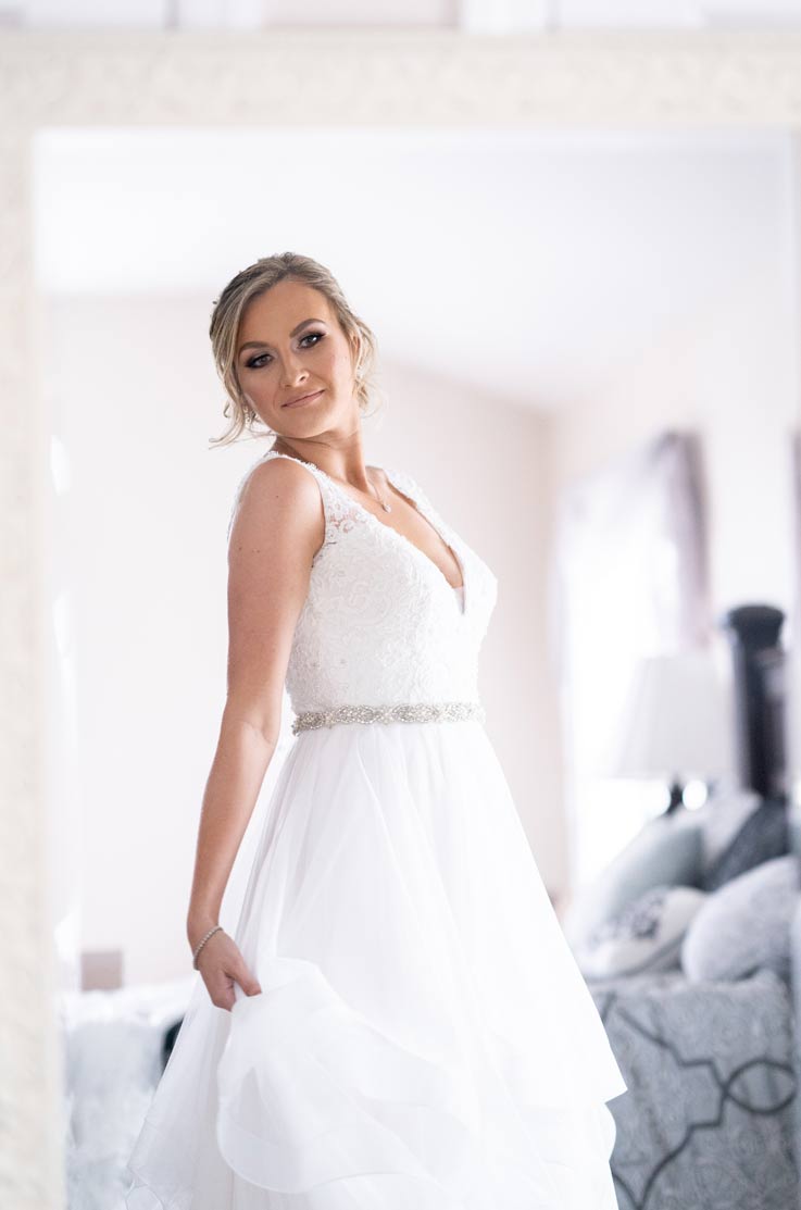 A bride in a tulle wedding dress holds her skirt and poses inside, with a bed in the background.