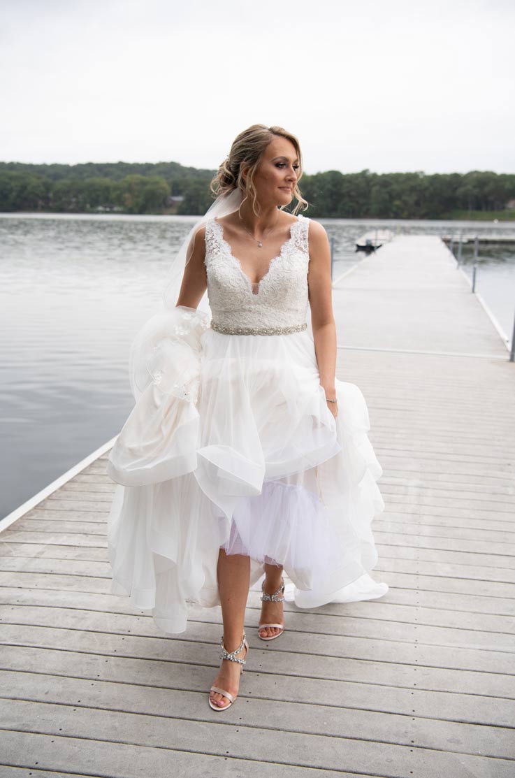 A bride in a tulle wedding dress holds her skirt up as she walks down a dock, with a lake and trees behind her.