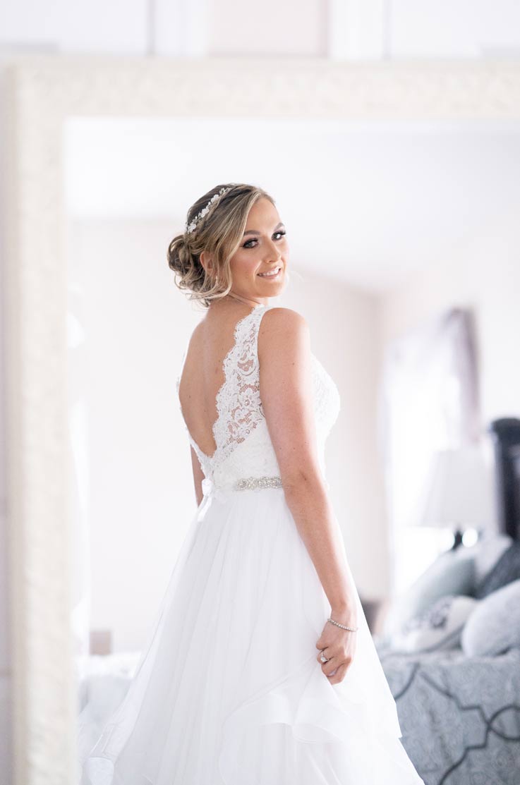 A smiling brie in a tulle wedding dress looks over her shoulder and poses inside, with a bed in the background.