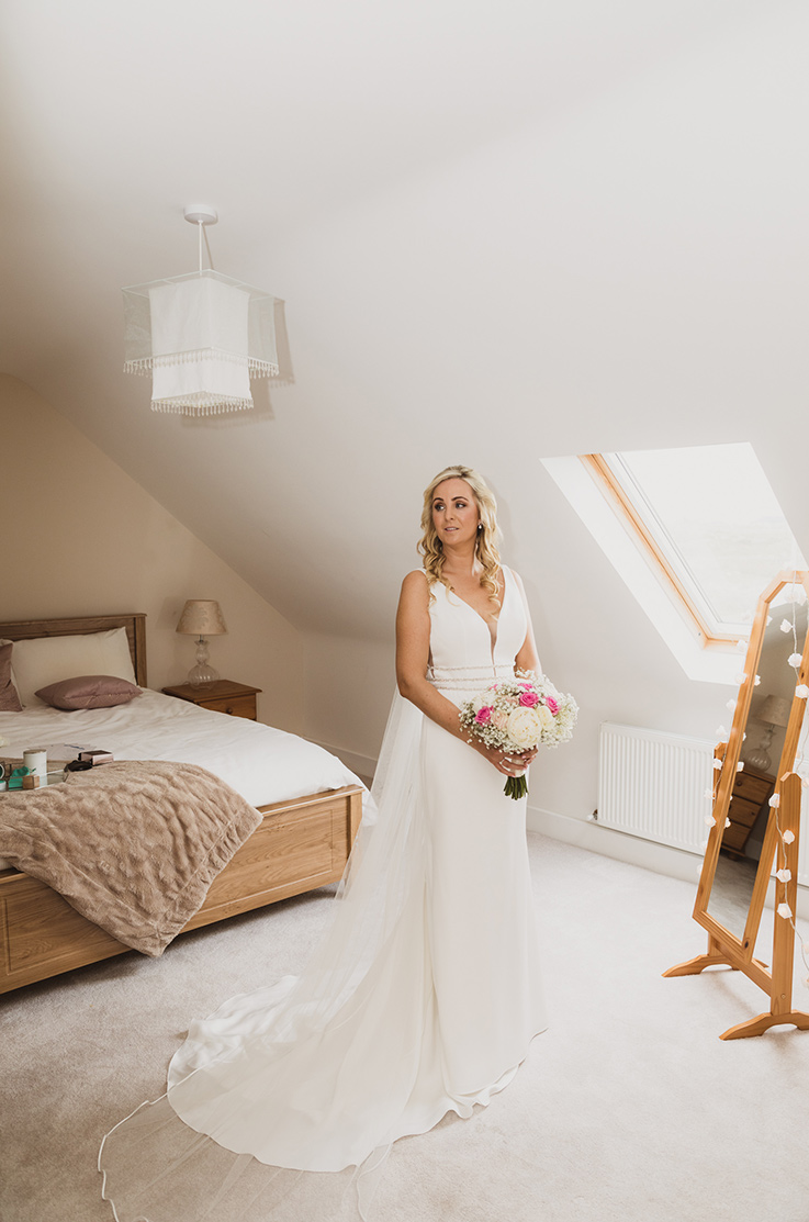 A bride in a V-neck wedding dress holding a bouquet poses indoors beside a mirror, with a window and a bed in the background.