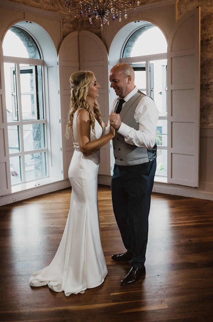 A brie in a sleeveless wedding dress and a groom in a suit jacket hold hands and smile at one another inside, with arched windows in the background and a chandelier above them.