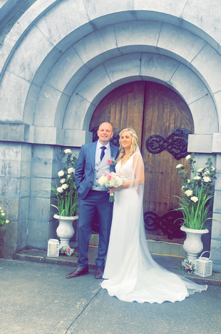 A bride in a V-neck wedding dress and veil, holding a bouquet, and a groom in a suit stand in front of a large arched stone entrance with large wooden doors.