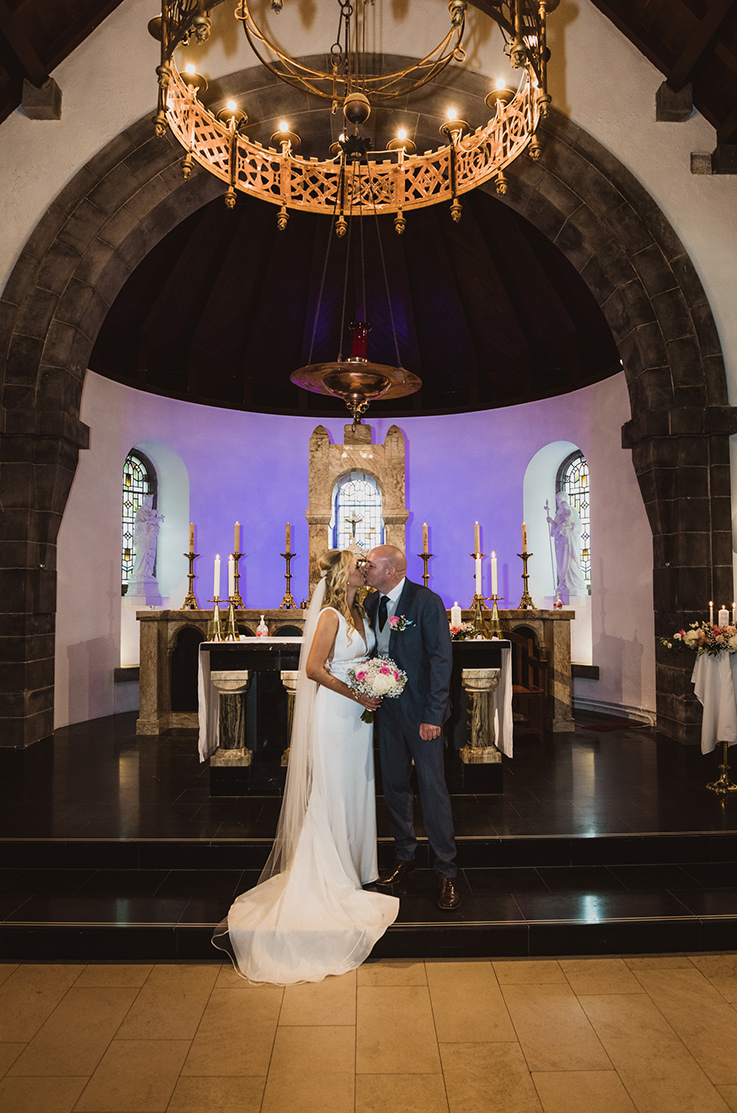 A bride in a V-neck wedding dress and veil, holding a bouquet, and a groom in a suit, kiss on the steps of an altar with a chandelier above them.